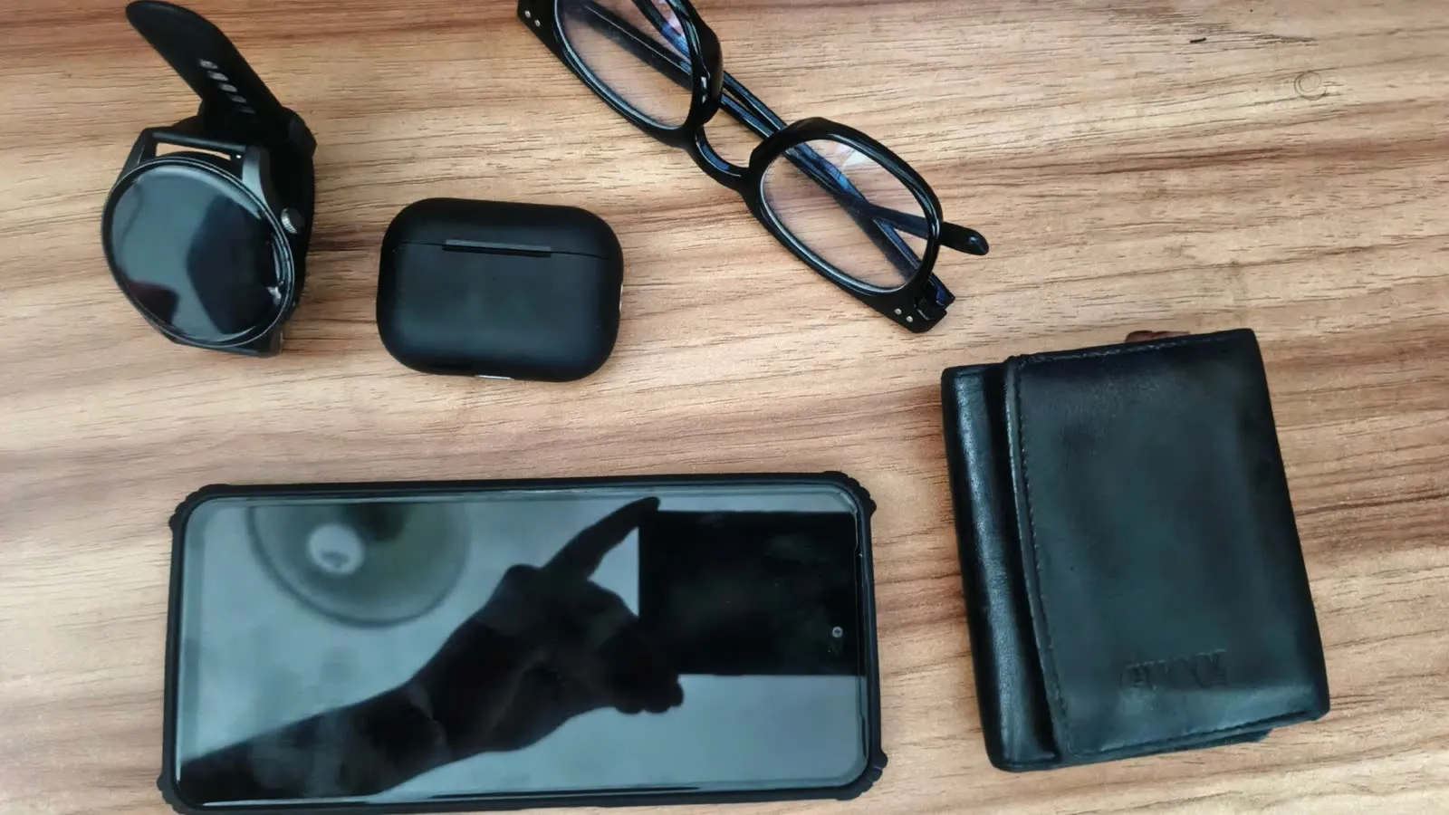 Estojo de fones sem fio, celular, óculos e carteira dispostos sobre uma mesa de madeira.