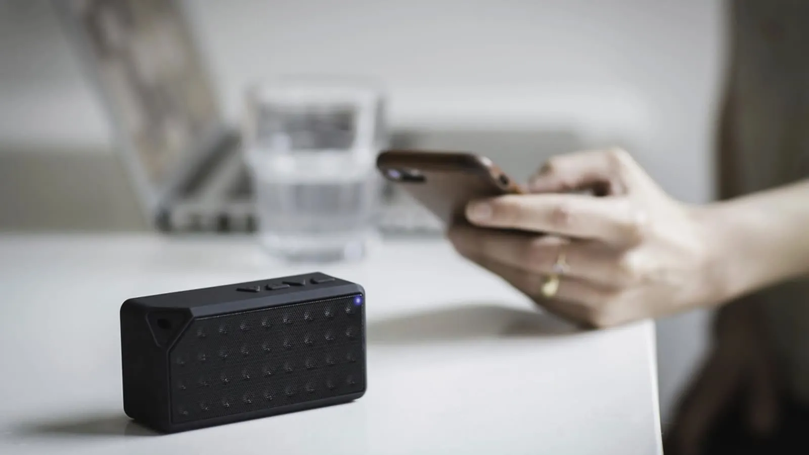 Caixa de som bluetooth compacta sobre a mesa, com uma pessoa mexendo no celular ao fundo.