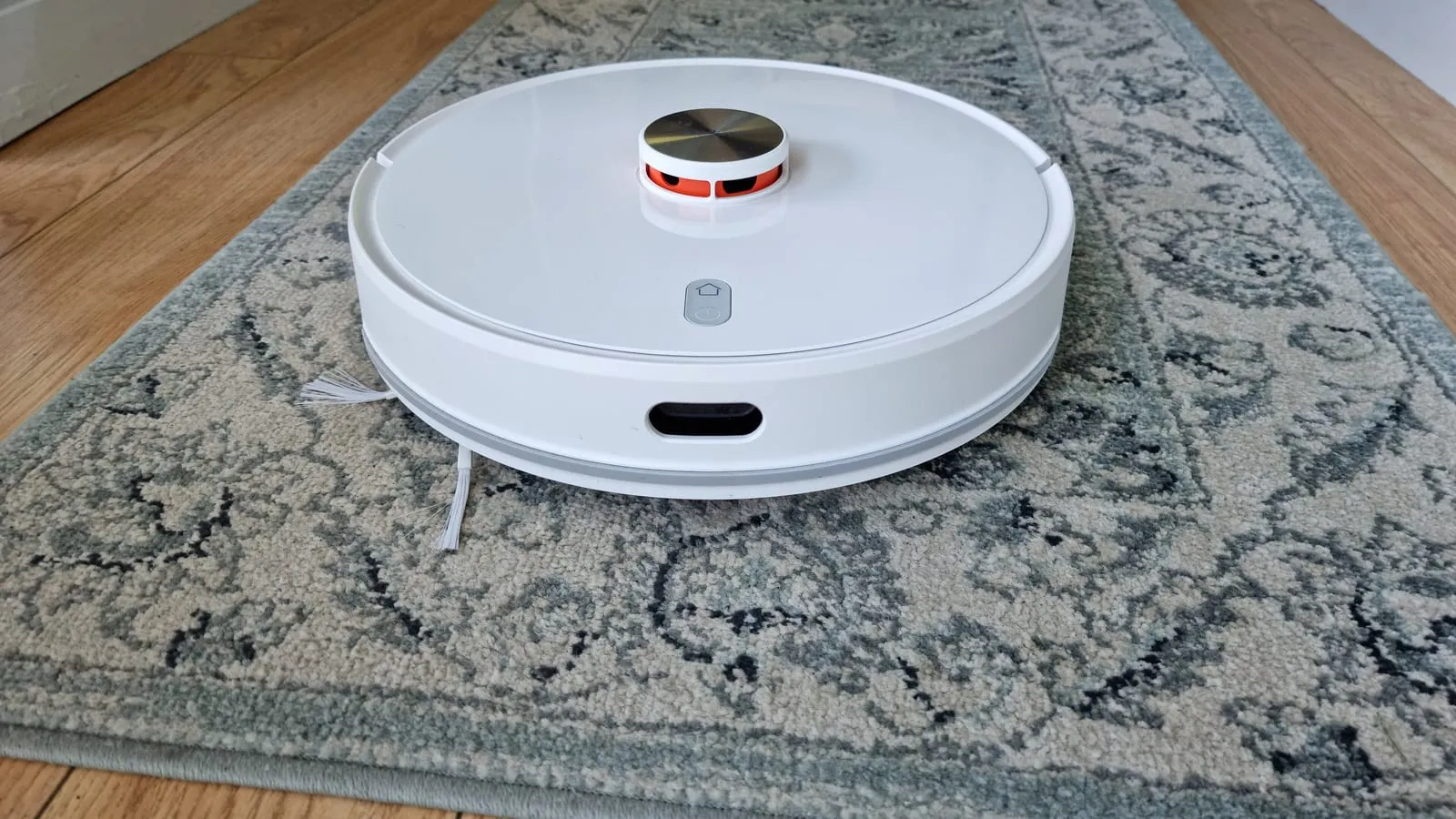 Robô aspirador branco com a torre do sensor a laser no topo, parado sobre um tapete estampado num corredor com piso de madeira.