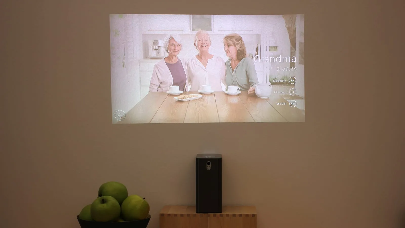 Projetor compacto sobre um caixote de madeira projetando uma imagem na parede de uma sala com a luz acesa, onde as cores aparecem desbotadas.