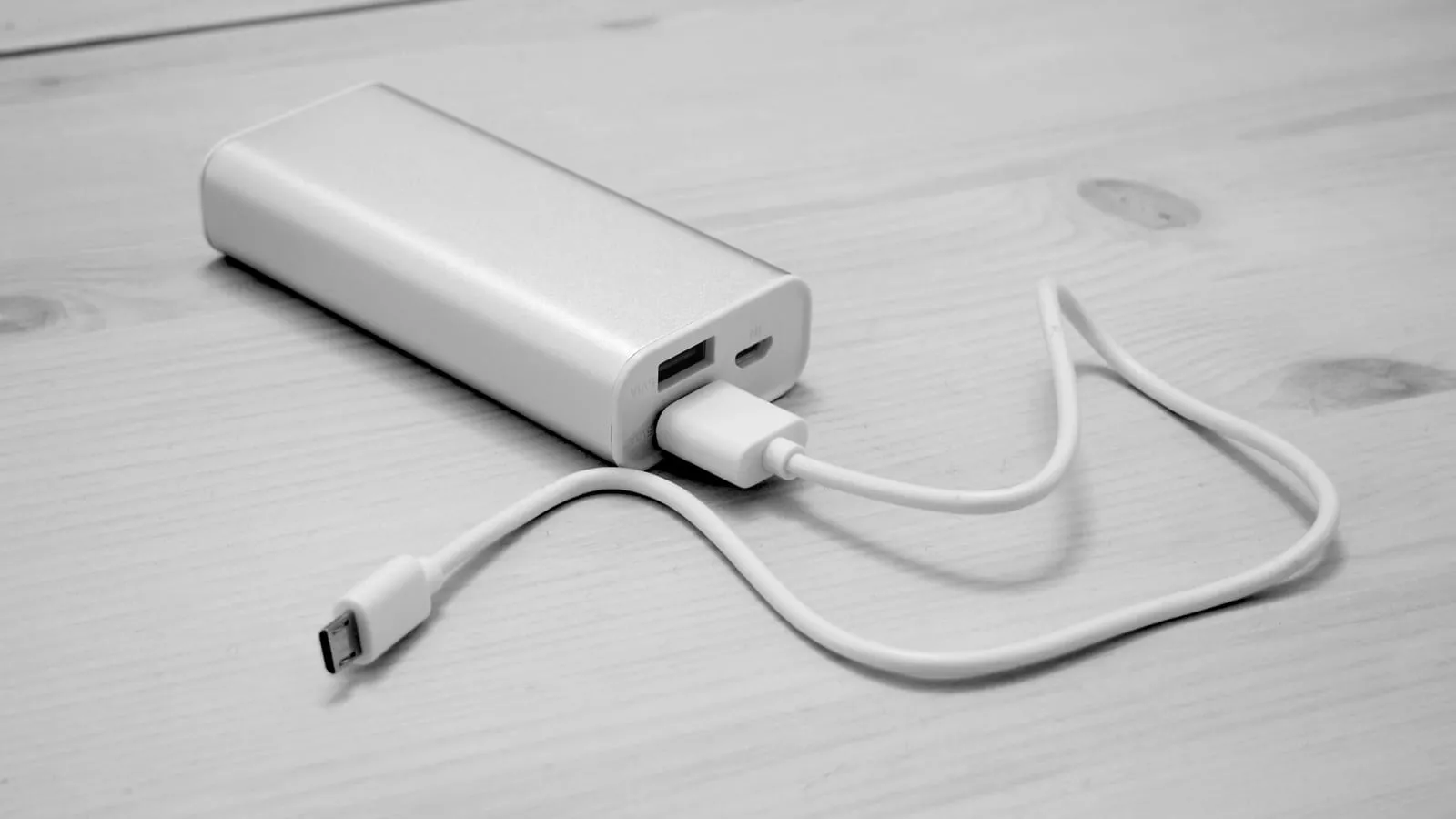 Power bank de alumínio sobre uma mesa de madeira clara, com cabo USB conectado.