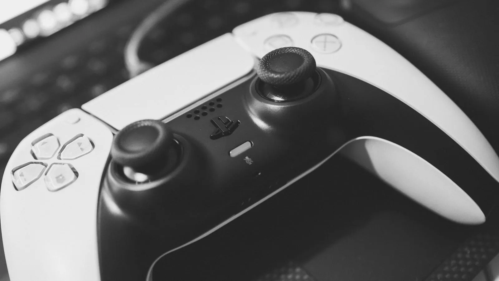 Controle DualSense do PlayStation 5 apoiado na mesa, com um teclado de computador desfocado ao fundo.